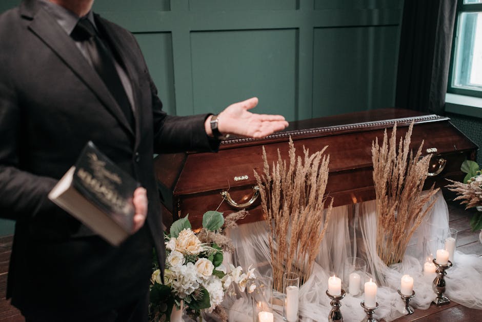 A person dressed in formal black attire is gesturing with one hand while holding a black book in the other, standing next to a wooden casket decorated with a floral arrangement. The arrangement featur