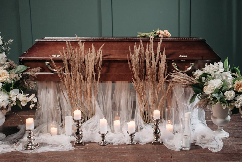 A wooden coffin is decorated with an arrangement of flowers, including white roses, cream-colored blossoms, and greenery, flanked by tall pampas grass stems. The display is set on a dark wooden floor