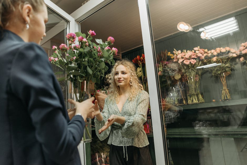 A woman with curly blonde hair and fair skin receives a bouquet of fresh pink roses from a florist through a glass door at Sally's Flower Shop. The florist, partially visible, has short hair and is we