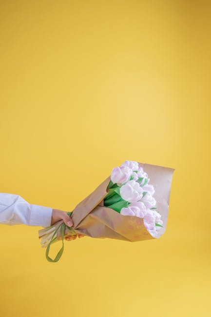 A person wearing a white shirt is holding a bouquet of fresh pink and white tulips wrapped in brown paper with green ribbon, positioned against a bright yellow background. The bouquet features vibrant