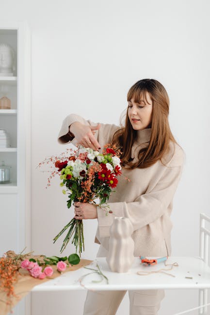 A woman with long, light brown hair and bangs wearing a beige sweater is arranging a vibrant floral bouquet in a bright, well-lit room. The bouquet features a variety of fresh flowers, including red,