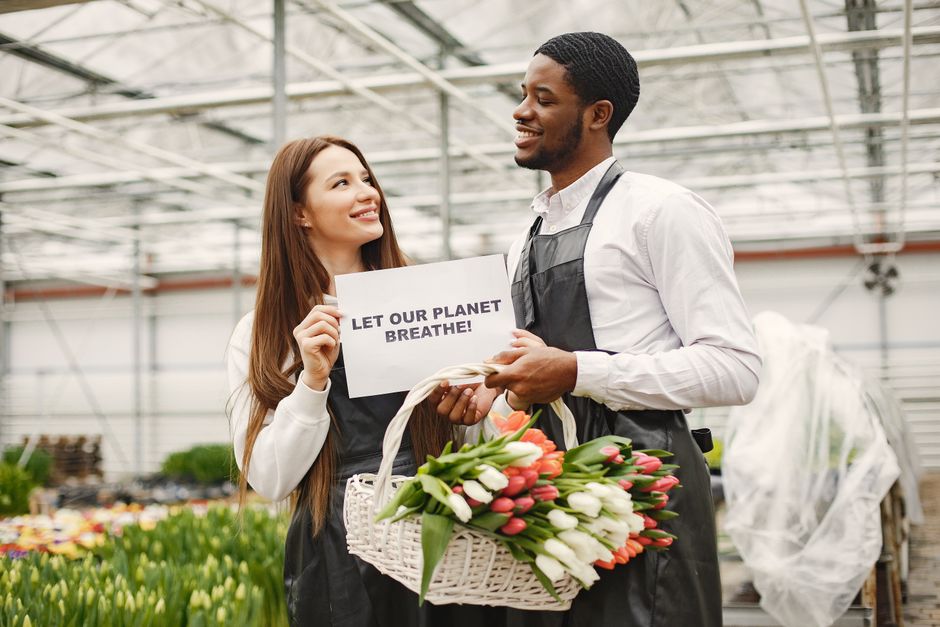 A young woman with long brown hair and a bright smile holds a small sign that reads 'LET OUR PLANET BREATHE!' while engaging in a conversation with a smiling man at a flower farm or greenhouse. The ma