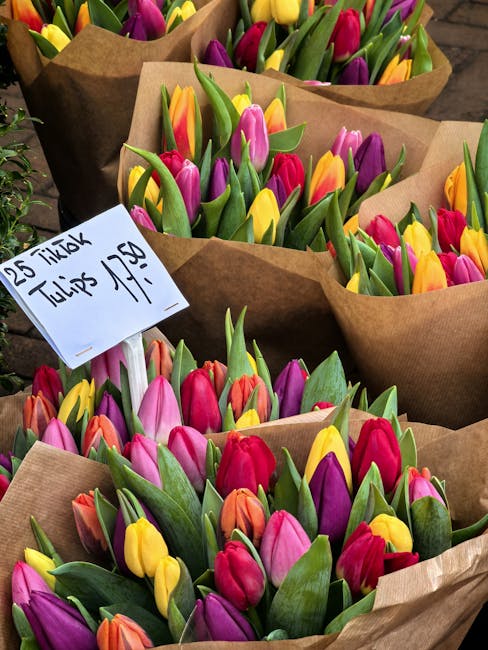 Close-up view of multiple bouquets of fresh tulips with vibrant pink, yellow, purple, and orange petals, wrapped in brown kraft paper, arranged on a flat surface. The tulips have green stems and leave