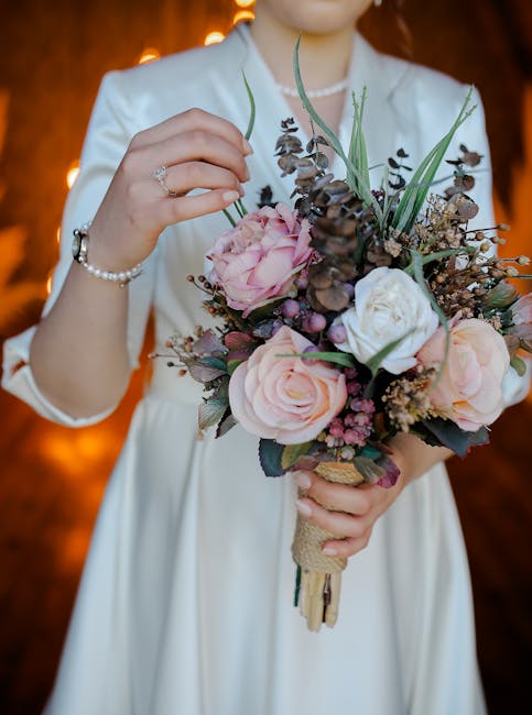 A person wearing a white satin dress is holding a floral bouquet with both hands, displaying the arrangement close to the chest. The bouquet features large, soft pink roses, white roses, and darker pu