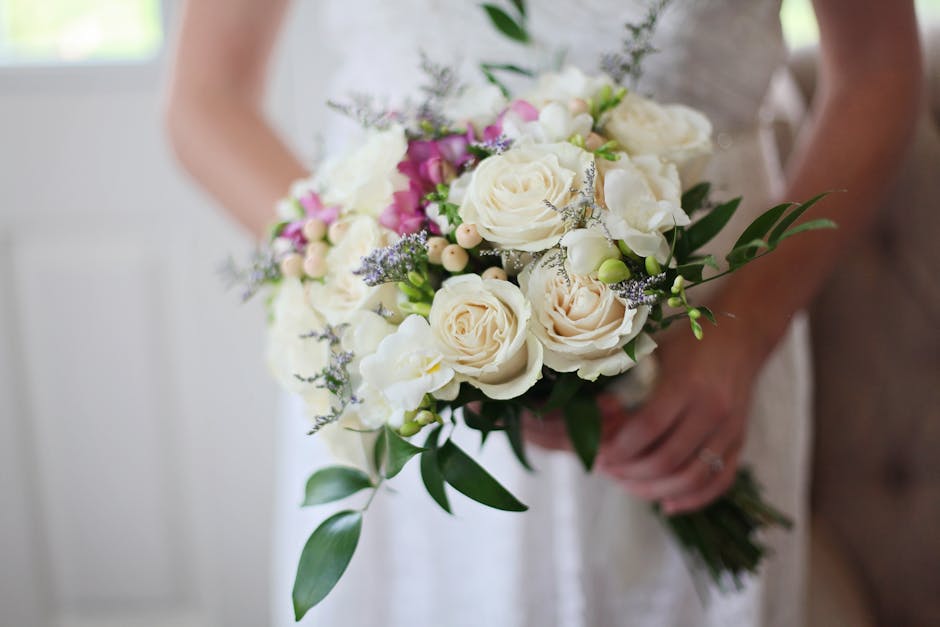 A person holding a bouquet of fresh floral arrangement featuring large, creamy white roses, smaller white flowers, and delicate sprigs of purple lavender, complemented by lush green foliage. The bouqu