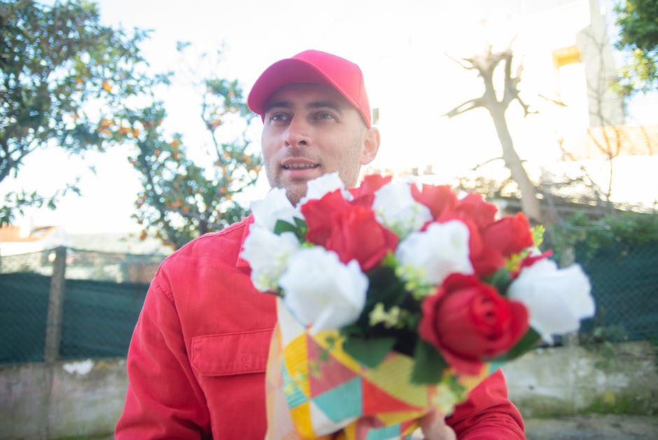 A delivery person wearing a red cap and matching red jacket holds a vibrant bouquet of fresh flowers outdoors, with red and white roses prominently visible, wrapped in colorful paper. The scene is bat