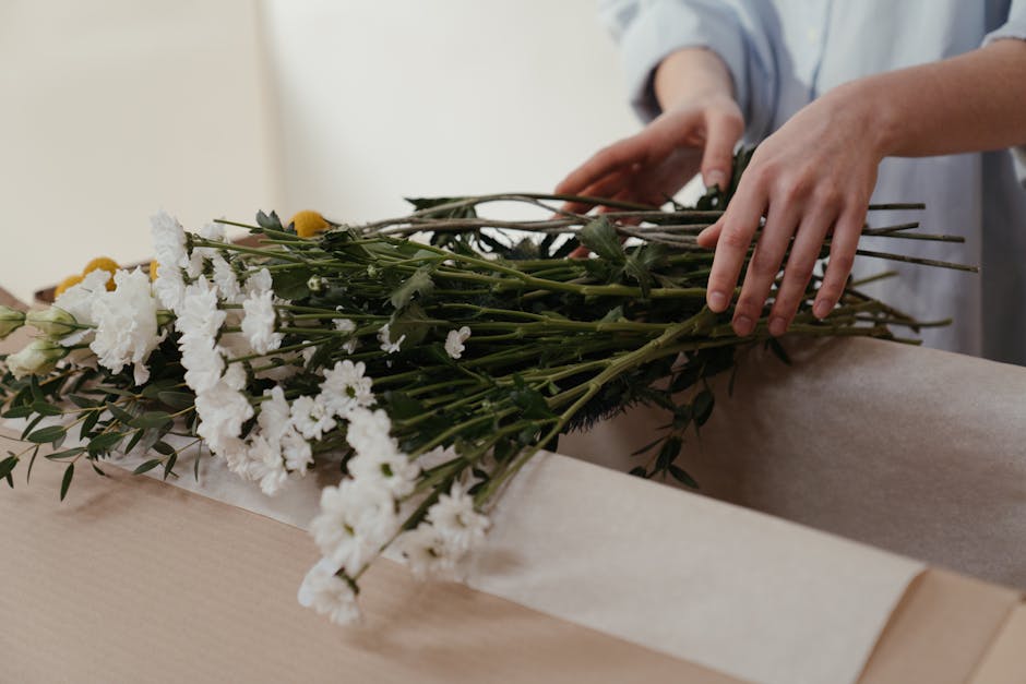 A person arranging a floral bouquet on a light wooden surface, featuring white carnations, yellow daisies, and green foliage wrapped in neutral-toned craft paper. Their hands are gently positioning th
