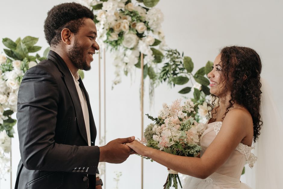 A bride and groom exchanging wedding vows indoors, with the bride holding a soft pastel-colored floral bouquet that includes roses, eucalyptus, and delicate filler flowers. The groom, dressed in a dar
