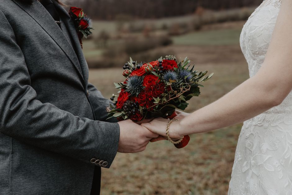 A bride and groom holding a vibrant bouquet of wedding flowers outdoors. The bouquet features deep red roses, blue thistles, dark berries, and leafy greenery, arranged in a natural, rounded style. The