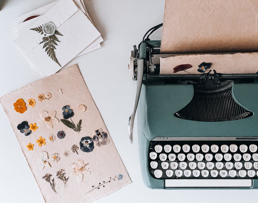 A vintage teal typewriter sits on a white surface with a sheet of handmade paper featuring an assortment of pressed flowers and leaves in various colours, including yellow, orange, blue, purple, and w