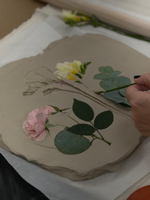 A person is carefully applying a yellow flower and sprigs of eucalyptus onto a piece of paper with pressed flowers, which appears to be part of creating a pressed flowers keepsake at home. The arrange