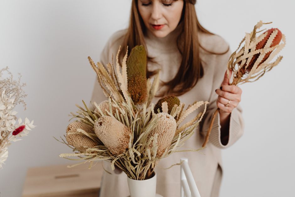A woman with long, reddish-brown hair arranging a bouquet of dried flowers in a white ceramic vase. The bouquet features beige and brown hues with textured, preserved protea and various dried grasses