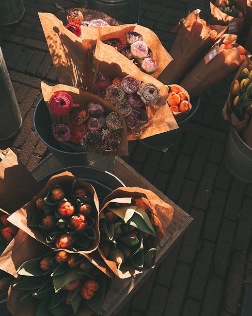 A variety of flower bouquets and floral arrangements displayed outdoors on wooden tables, illuminated by natural sunlight. The bouquets are wrapped in brown kraft paper and arranged in metal buckets a