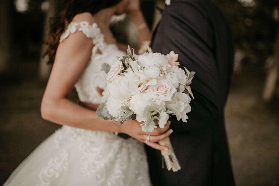 Close-up of a bride and groom holding a floral bouquet during a wedding. The bouquet features soft pink and white roses, pale green and white hydrangeas, and dusty miller leaves, arranged in a rounded