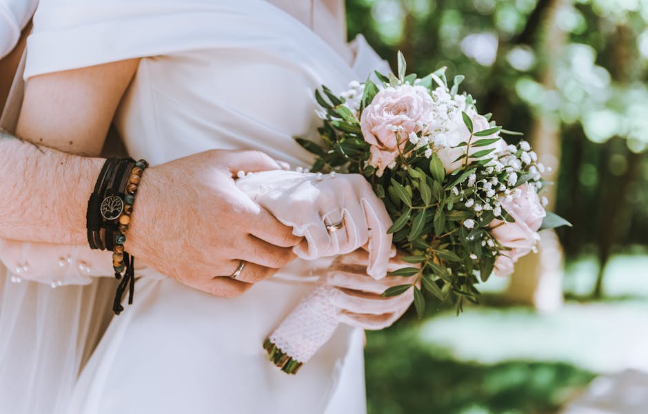 Close-up of a bride holding a bouquet of fresh flowers, including pale pink roses, white baby's breath, and green foliage, wrapped in light pink and white patterned paper with a matching ribbon. The b